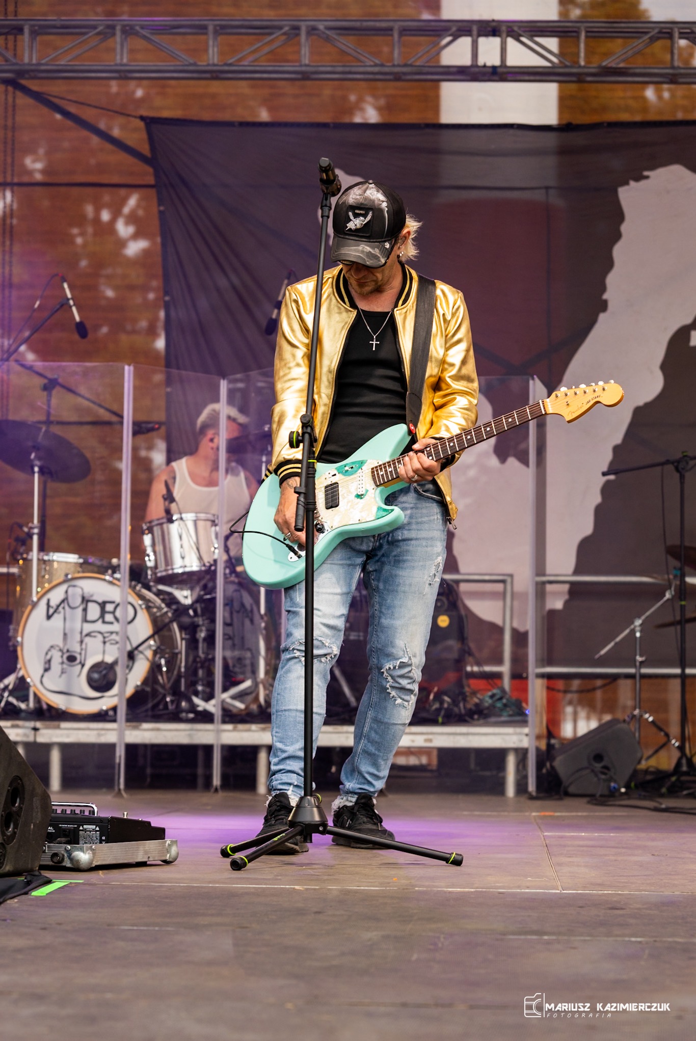 Muzyk w złotej kurtce i czapce z daszkiem gra na jasnoniebieskiej gitarze elektrycznej na scenie, w tle perkusista.
