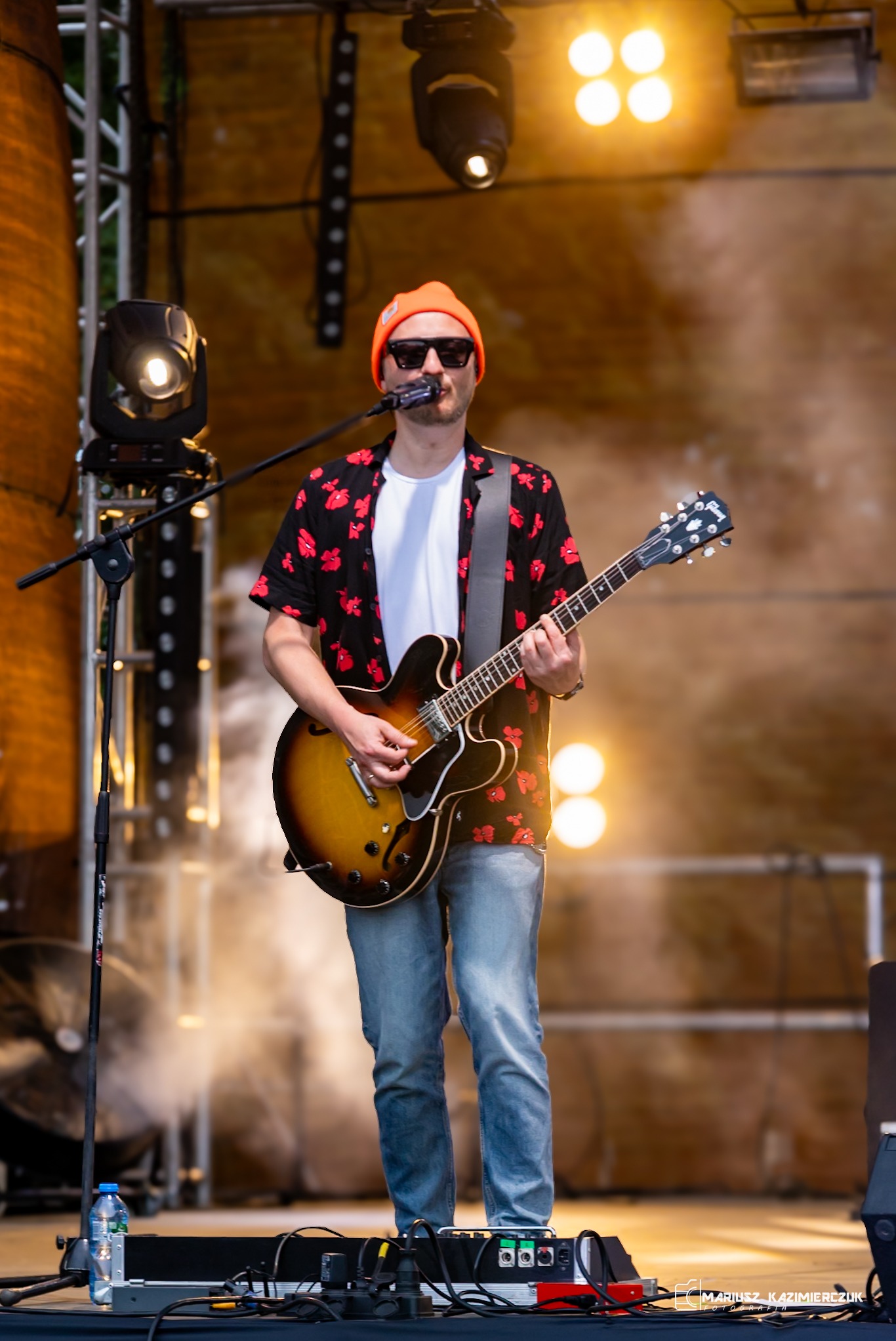 Muzyk grający na gitarze elektrycznej na scenie, ubrany w czarną koszulę w czerwone kwiaty i pomarańczową czapkę, oświetlony reflektorami.