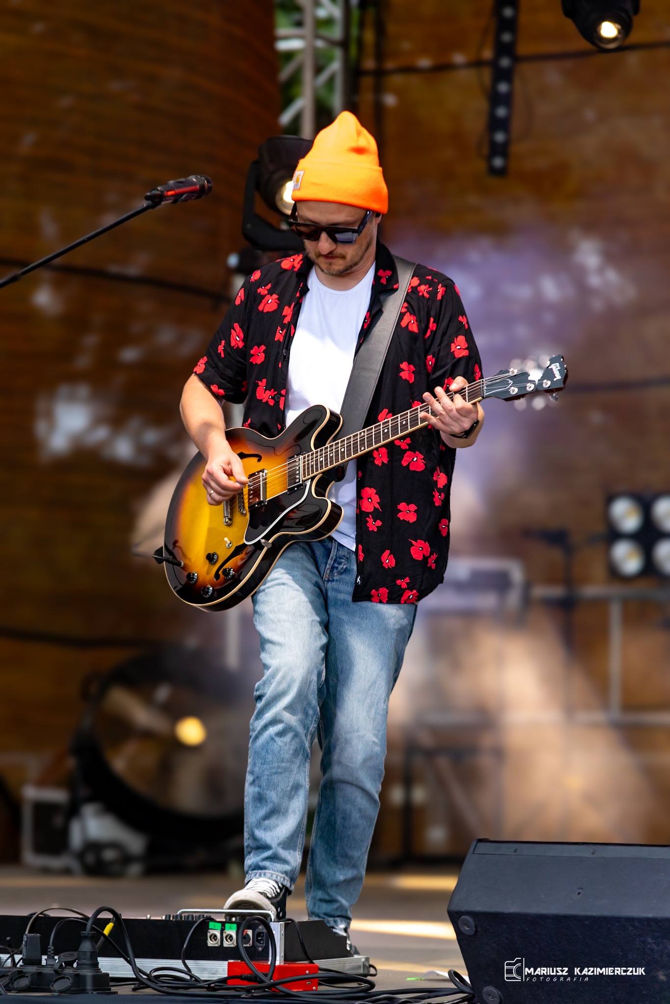 Muzyk grający na gitarze elektrycznej na scenie, ubrany w pomarańczową czapkę i koszulę w kwiaty, z widocznym mikrofonem i sprzętem nagłośnieniowym.