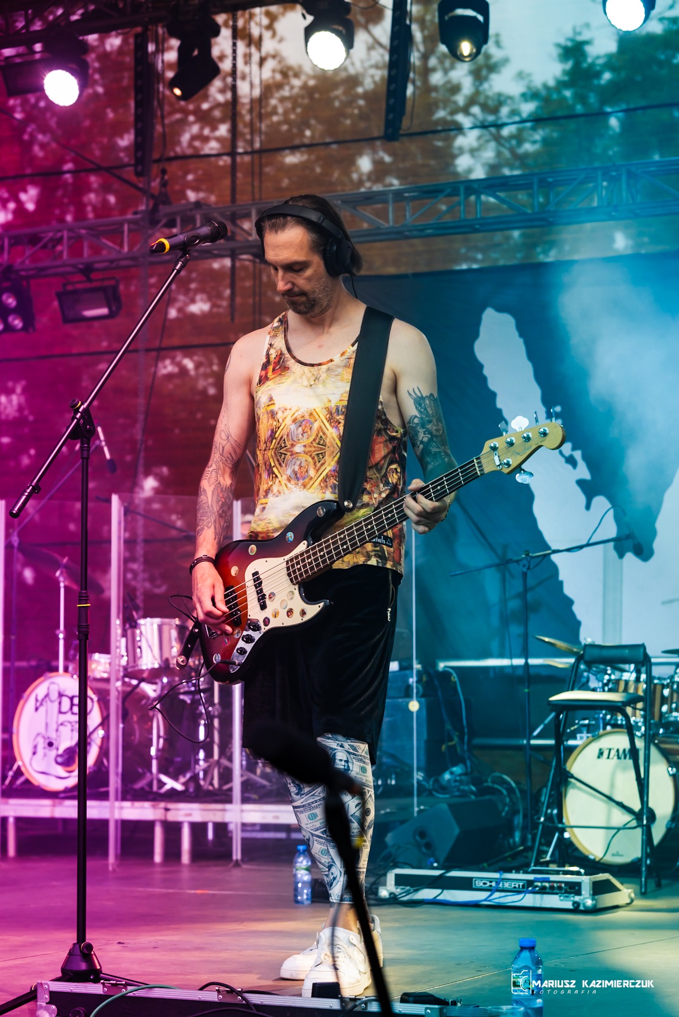 Basista z tatuażami gra na scenie, ubrany w koszulkę z nadrukiem i legginsy z motywem banknotów, w tle perkusja i oświetlenie sceniczne.