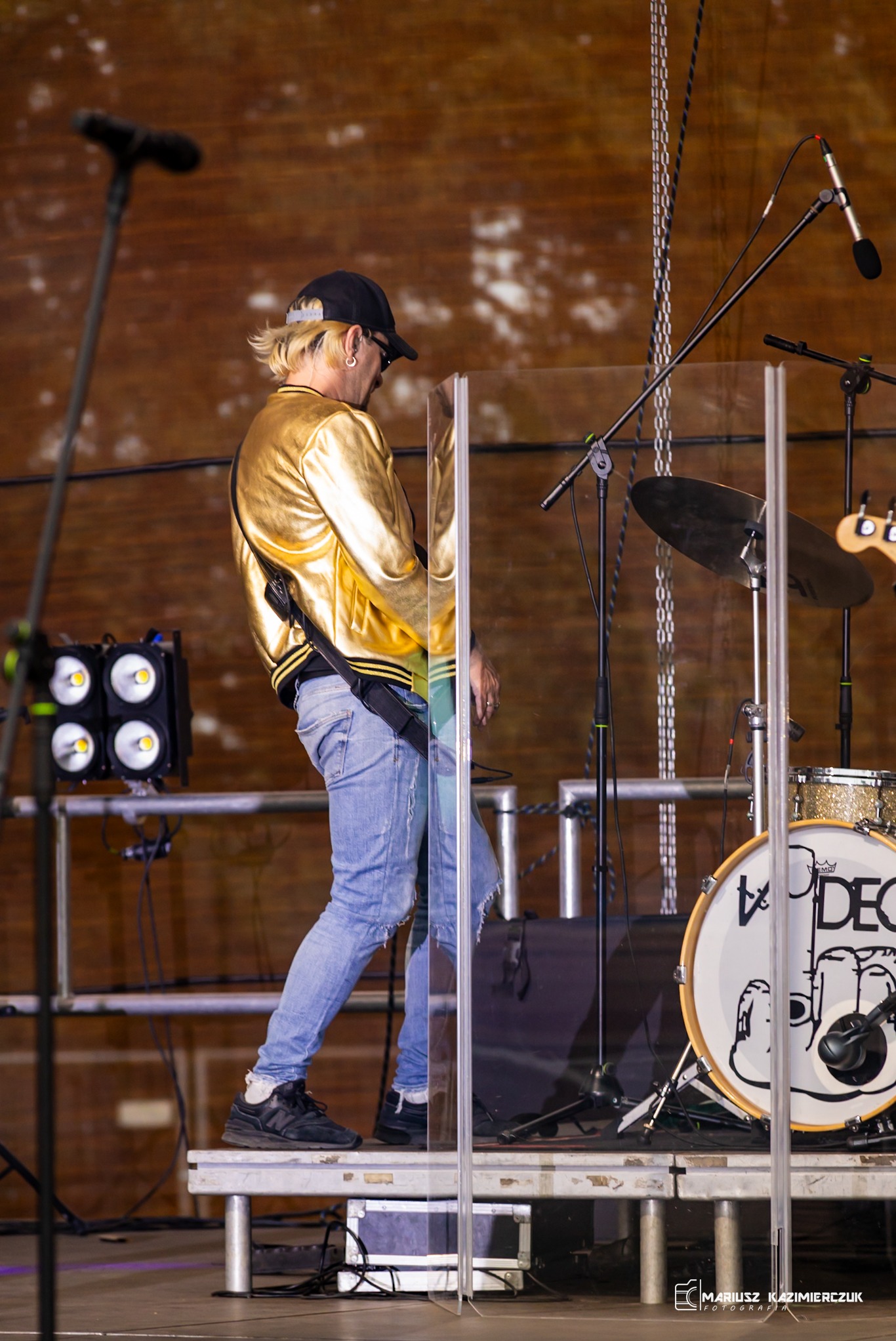 Muzyk w złotej kurtce i czapce bejsbolowej gra na gitarze na scenie, widoczny boczny profil, instrumenty perkusyjne i oświetlenie sceniczne w tle.