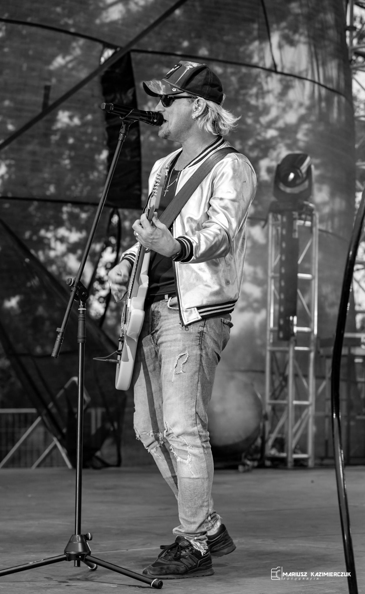 Czarno-białe zdjęcie muzyka z blond włosami w srebrnej kurtce grającego na białej gitarze elektrycznej na scenie, z mikrofonem i sprzętem oświetleniowym w tle.