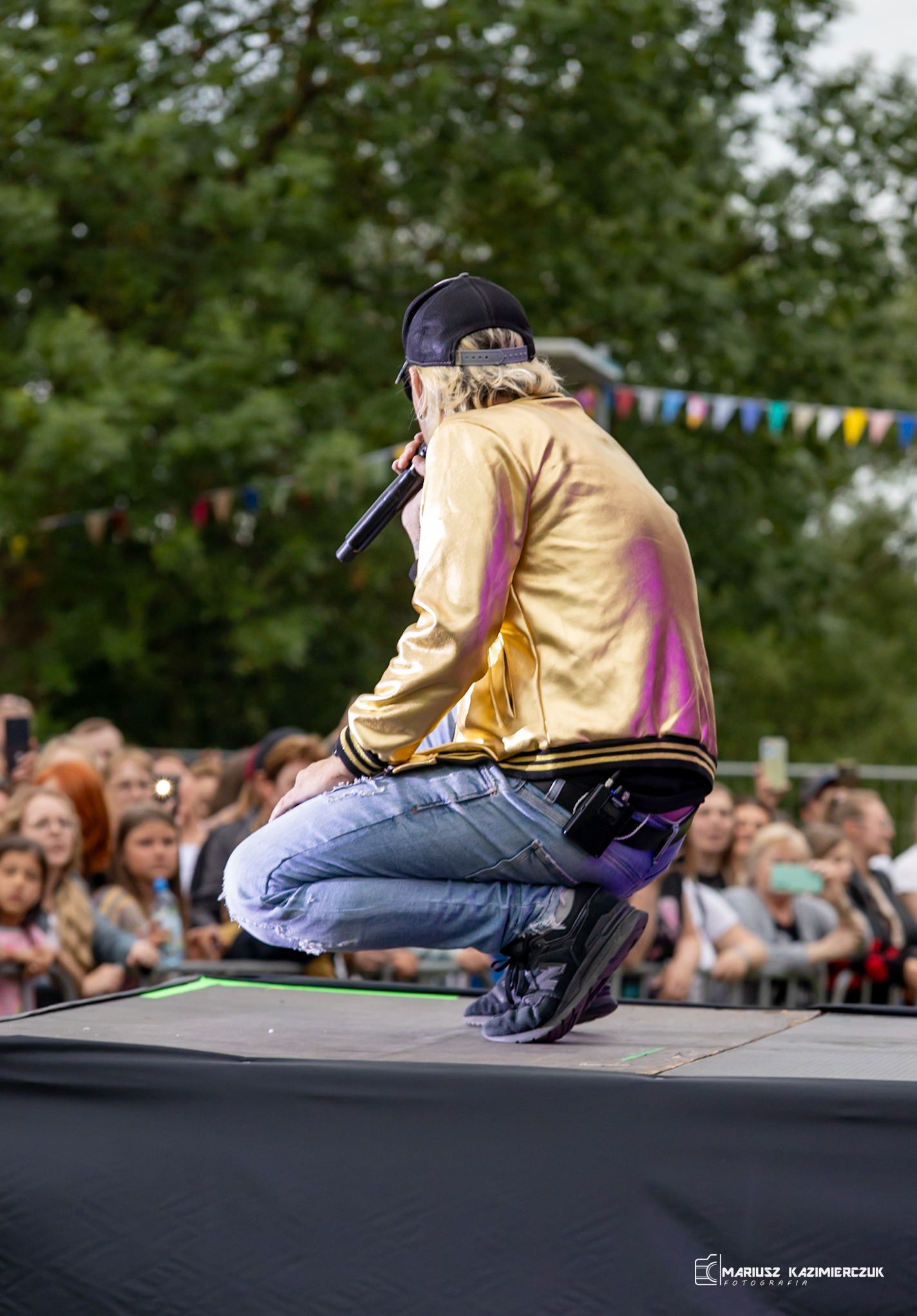 Piosenkarz w złotej kurtce i czapce, kuca na scenie podczas występu, trzymając mikrofon, z widoczną publicznością w tle.
