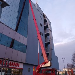 podnośniki koszowe Liftpol - Czerwony podnośnik koszowy na tle nowoczesnego budynku biurowego z przeszkloną fasadą, widoczny kosz z pracownikiem na wysokości, obok budynek z balkonami i szyld sklepu Rossmann