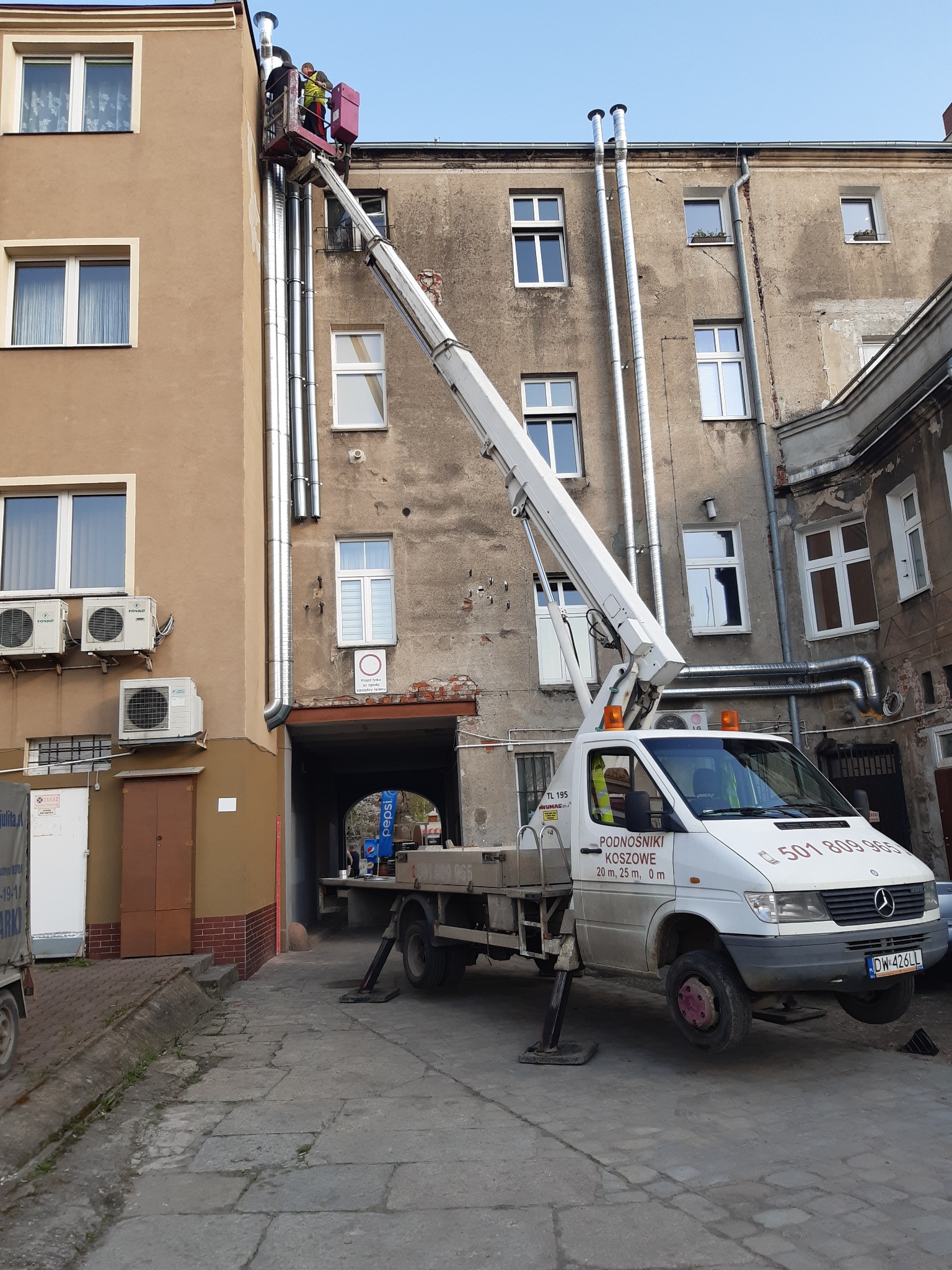 Pracownicy w koszu podnośnika montują rury spustowe na elewacji budynku, widoczny biały samochód z napisem 'Podnośniki Koszowe' i numerem telefonu.