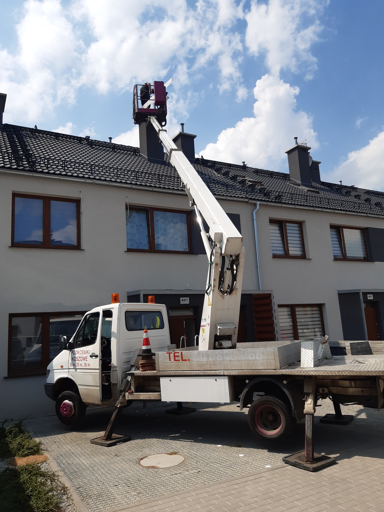 Biały podnośnik koszowy na pojeździe ustawiony przed budynkiem mieszkalnym z ciemną dachówką, kosz podnośnika na wysokości dachu, błękitne niebo w tle.