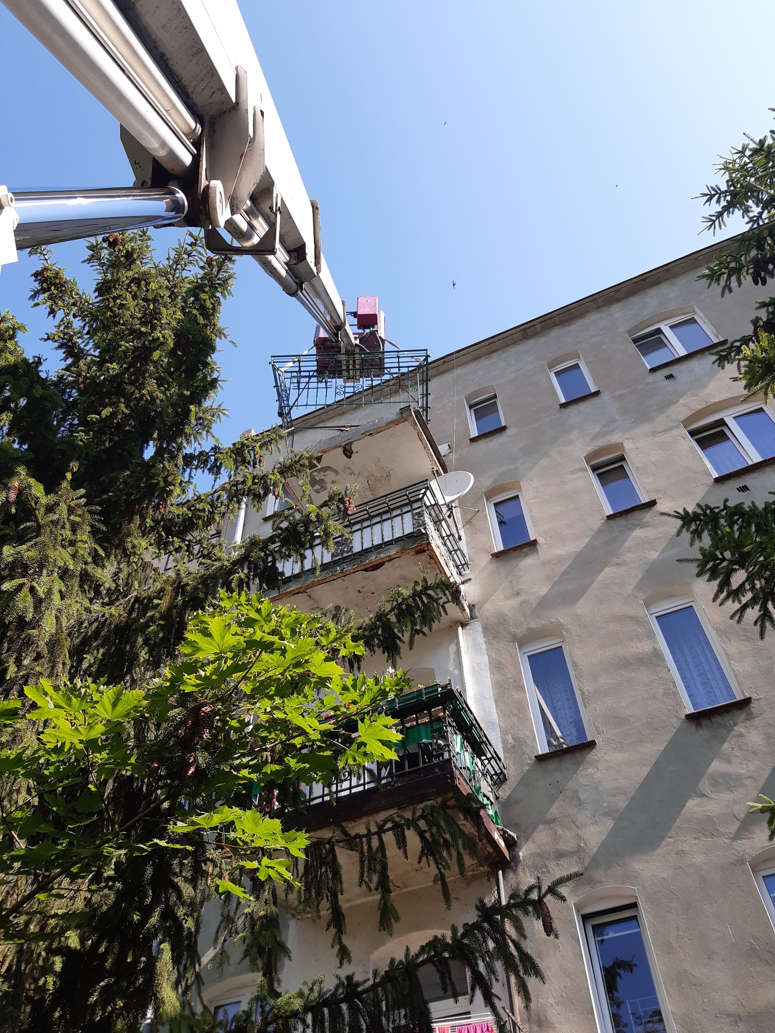 Podnośnik koszowy z koszem na wysokości balkonu w starym budynku, widok z dołu, drzewa zasłaniają część fasady.