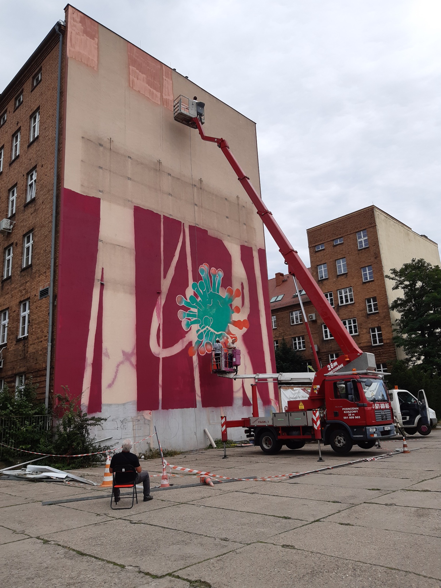 Malowanie muralu z podnośnika koszowego na ścianie budynku, widoczne fragmenty wzoru, czerwone pasy i zielony motyw graficzny, zabezpieczenia na ziemi.