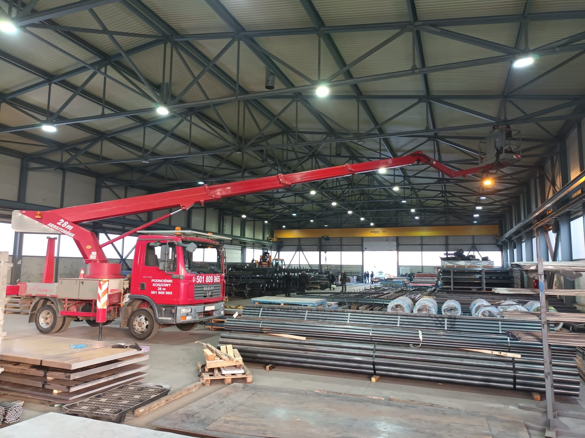Czerwony podnośnik koszowy na samochodzie ciężarowym z wysięgnikiem podniesionym wewnątrz hali magazynowej, wśród rur i metalowych elementów konstrukcyjnych.