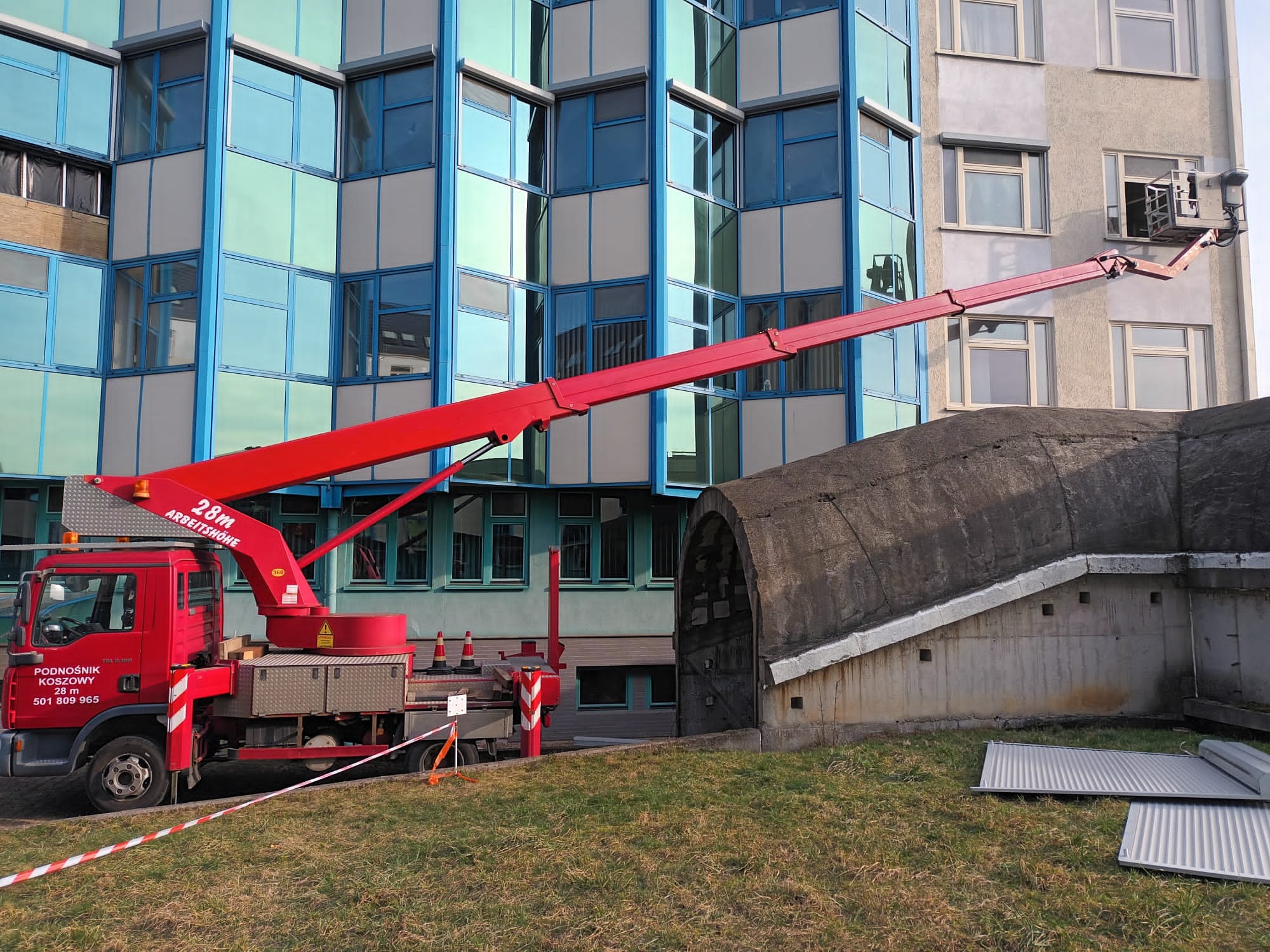 Czerwony podnośnik koszowy ustawiony przy budynku z niebieskimi oknami, kosz podnośnika przy oknie na wyższej kondygnacji, obok widoczny betonowy schron.