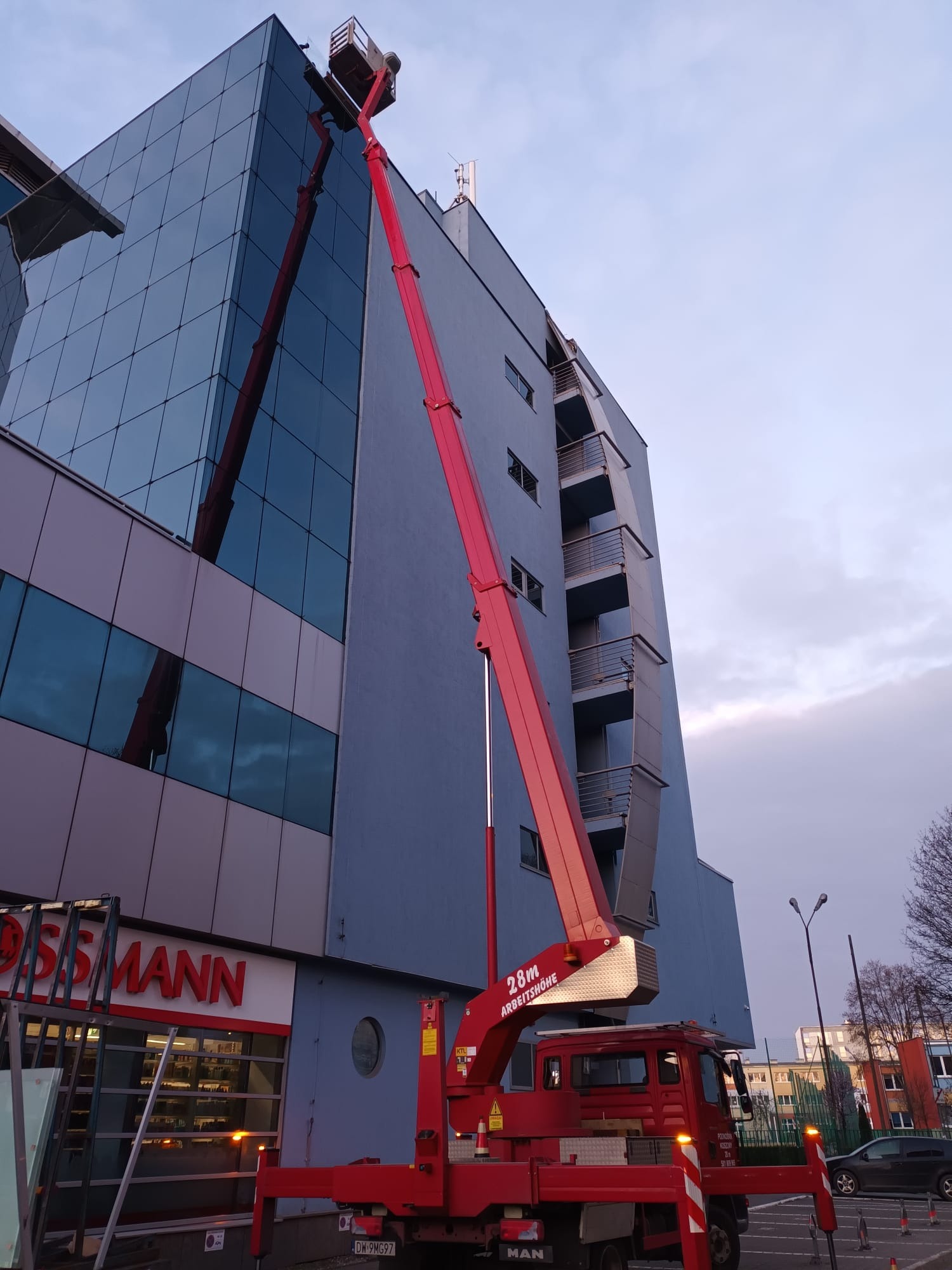 Czerwony podnośnik koszowy na tle nowoczesnego budynku biurowego z przeszkloną fasadą, widoczny kosz z pracownikiem na wysokości, obok budynek z balkonami i szyld sklepu Rossmann