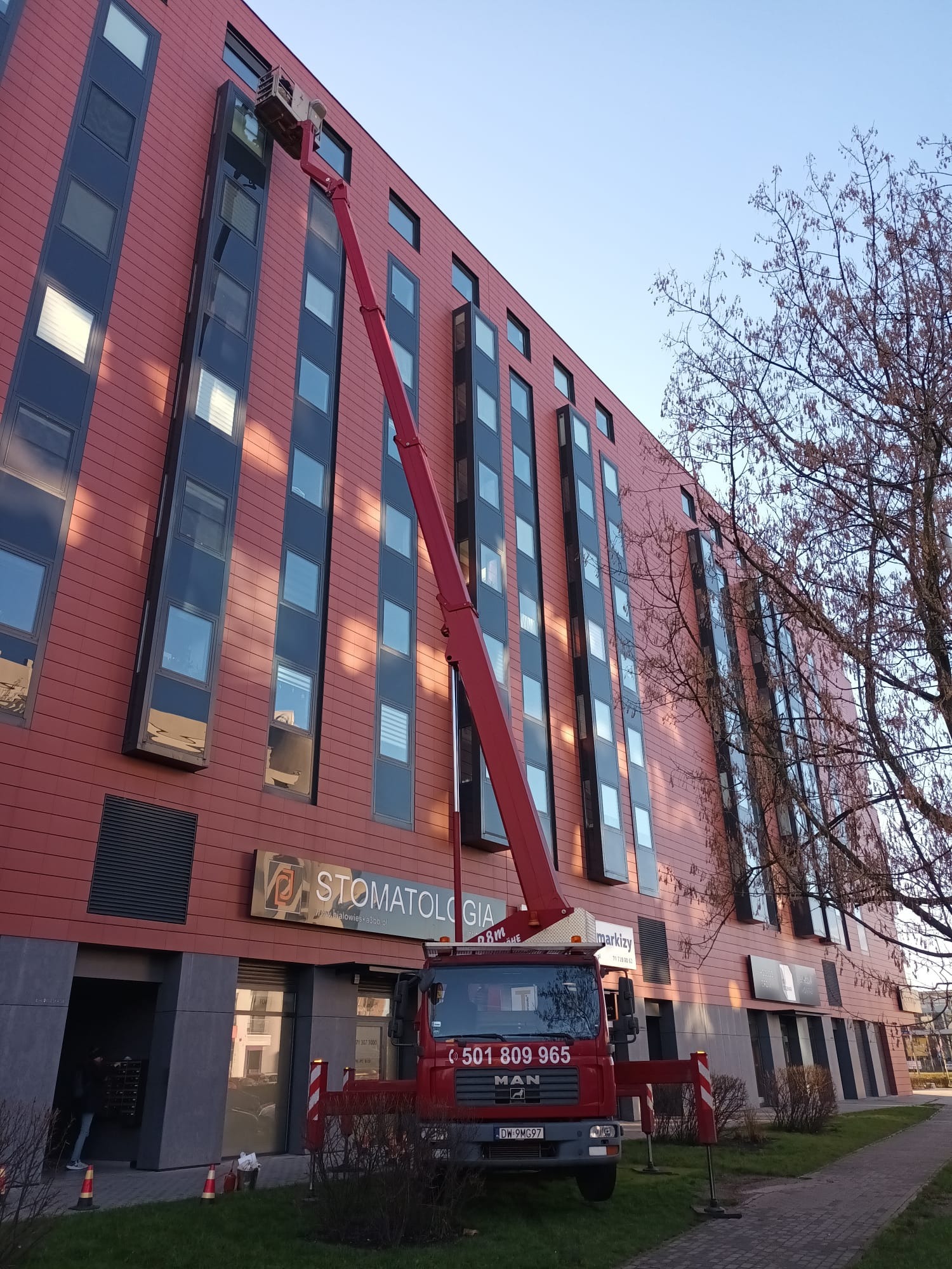 Czerwony podnośnik koszowy MAN na tle nowoczesnego budynku z licznymi oknami, przed wejściem do gabinetu stomatologicznego, z widoczną nazwą firmy i numerem telefonu na kabinie.