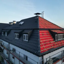 Budynek z częściowo zdjętym, starym pokryciem i widoczną czerwoną membraną oraz łatami, pozostała część dachu pokryta ciemną dachówką, rusztowania przy ścianie budynku.