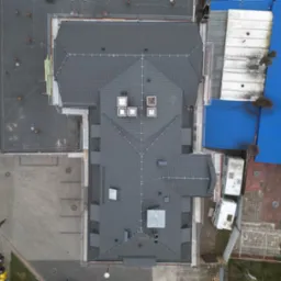 Widok z góry na budynek z nowym, ciemnoszarym pokryciem dachowym, widoczne kominy wentylacyjne, fragment rusztowania i niebieski dach sąsiedniego budynku.