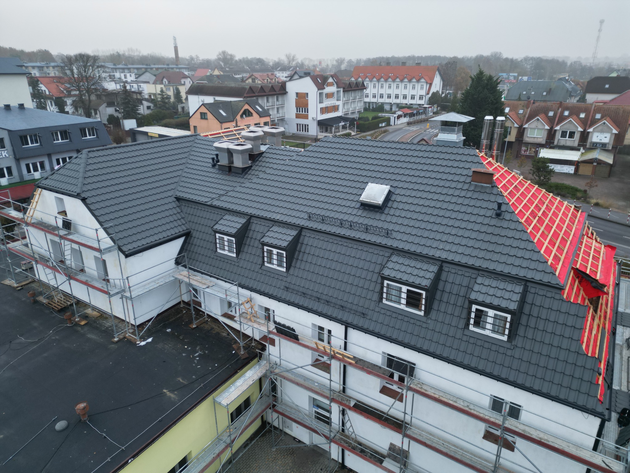 Widok z góry na budynek z częściowo zdjętym i nowym pokryciem, w trakcie prac dekarskich z użyciem rusztowań, na tle zabudowy miejskiej w pochmurny dzień.