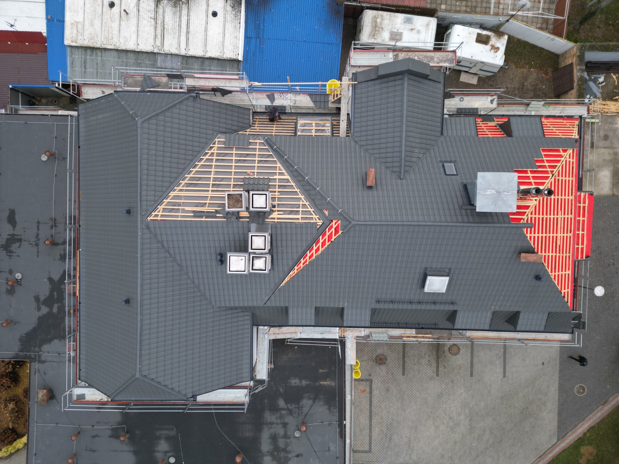 Widok z góry na budynek z częściowo zdjętym i częściowo nowym, grafitowym pokryciem. Na fragmencie widoczna drewniana konstrukcja dachu, a na innym czerwone łaty pod nowe pokrycie. W tle sąsiednie...