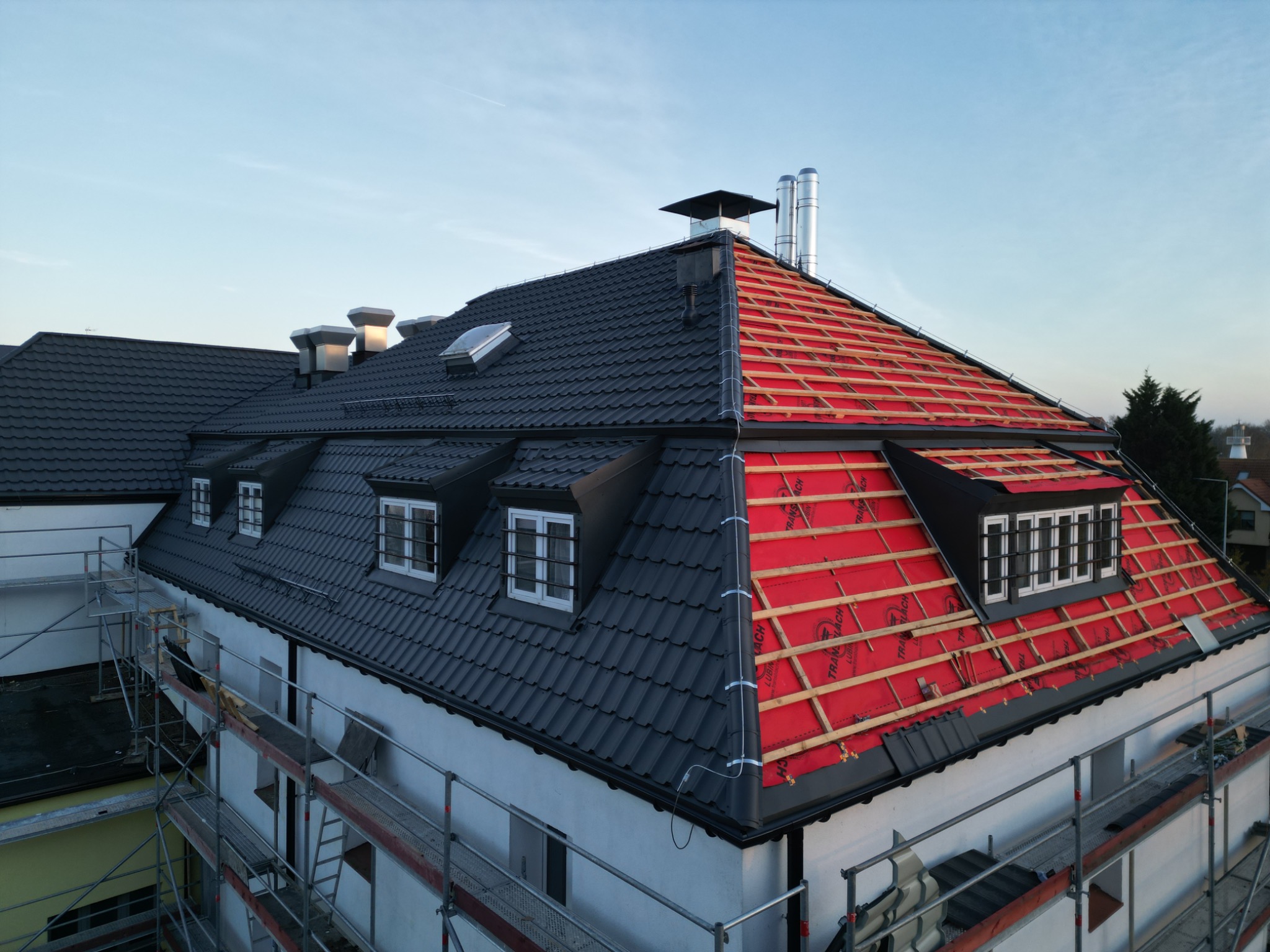 Budynek z częściowo zdjętym, starym pokryciem i widoczną czerwoną membraną oraz łatami, pozostała część dachu pokryta ciemną dachówką, rusztowania przy ścianie budynku.
