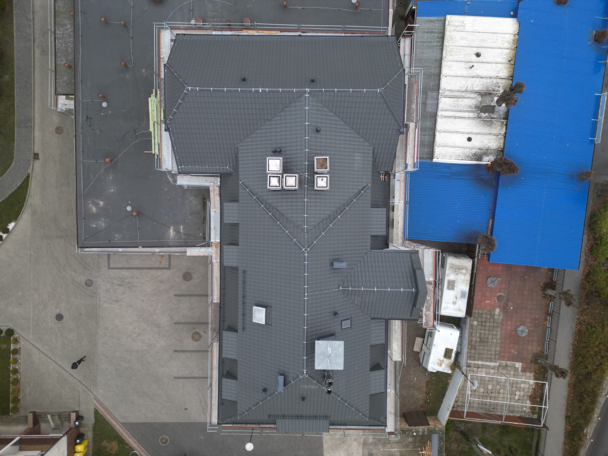 Widok z góry na budynek z nowym, ciemnoszarym pokryciem dachowym, widoczne kominy wentylacyjne, fragment rusztowania i niebieski dach sąsiedniego budynku.