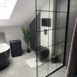 Nowoczesna łazienka na poddaszu z kabiną prysznicową typu walk-in z czarnymi szprosami, marmurowymi płytkami, czarną armaturą i dodatkami w odcieniach szarości i złota, oświetlona światłem...