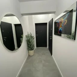 Nowoczesny korytarz z szarymi płytkami podłogowymi, owalnym lustrem, rośliną w beżowej doniczce i obrazem krowy w lustrzanej ramie.