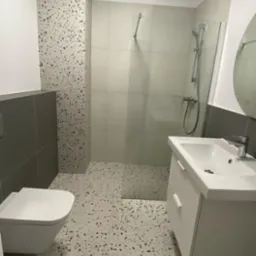 Nowoczesna łazienka z kabiną prysznicową ze szklaną ścianką, białą umywalką z szafką, podwieszaną toaletą i podłogą z płytek lastryko.