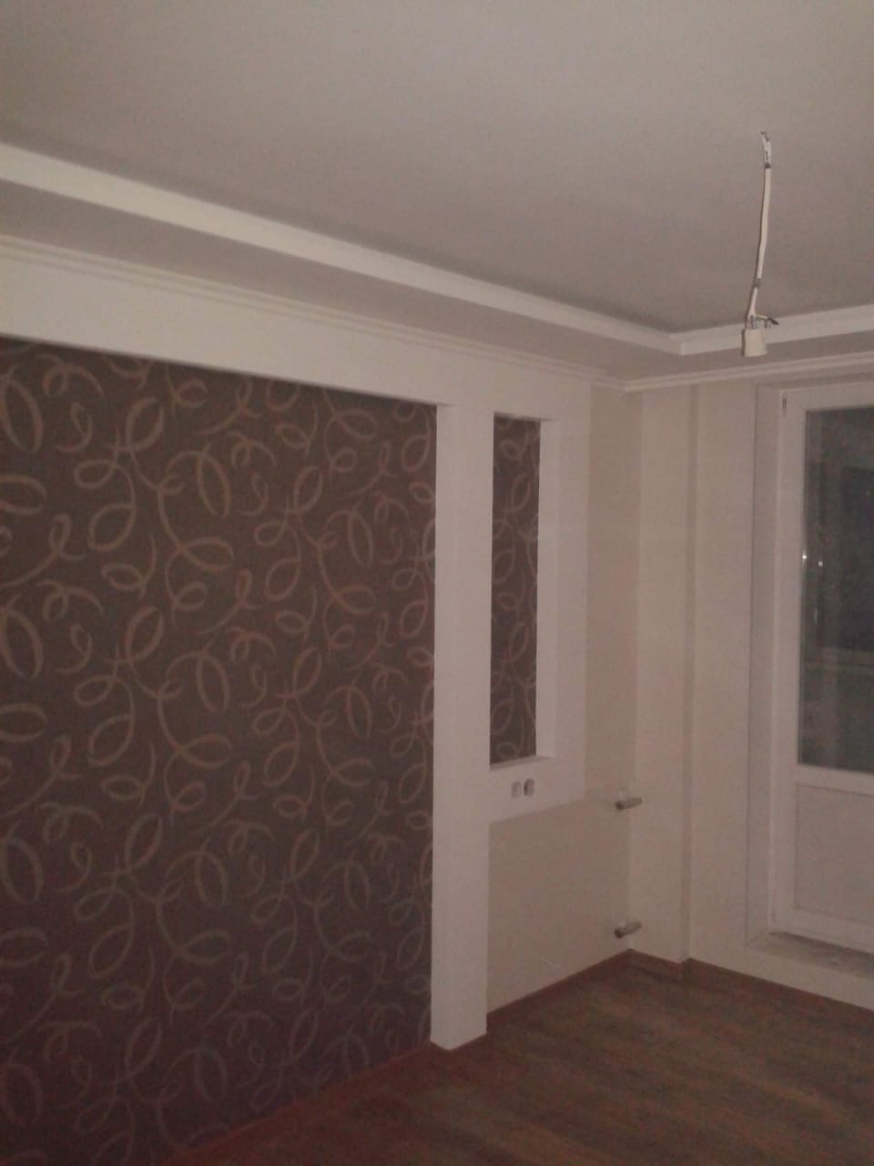 Pokój po remoncie z brązową tapetą w ornamenty, białe listwy przysufitowe, drewniana podłoga, wystający kabel z sufitu.