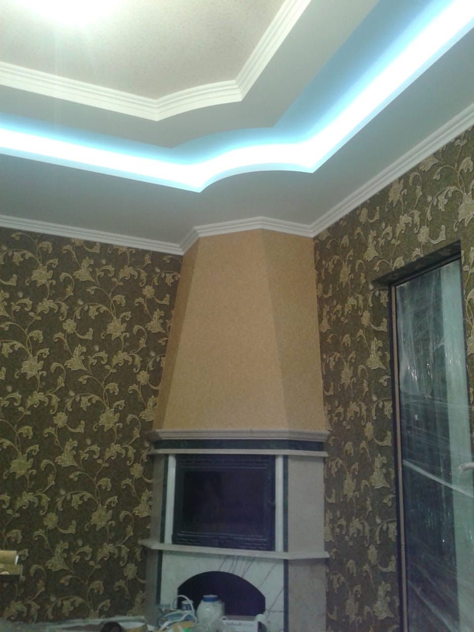 Wnętrze pokoju z kominkiem wykończonym marmurem, tapetą w kwiaty i podwieszanym sufitem z oświetleniem LED.