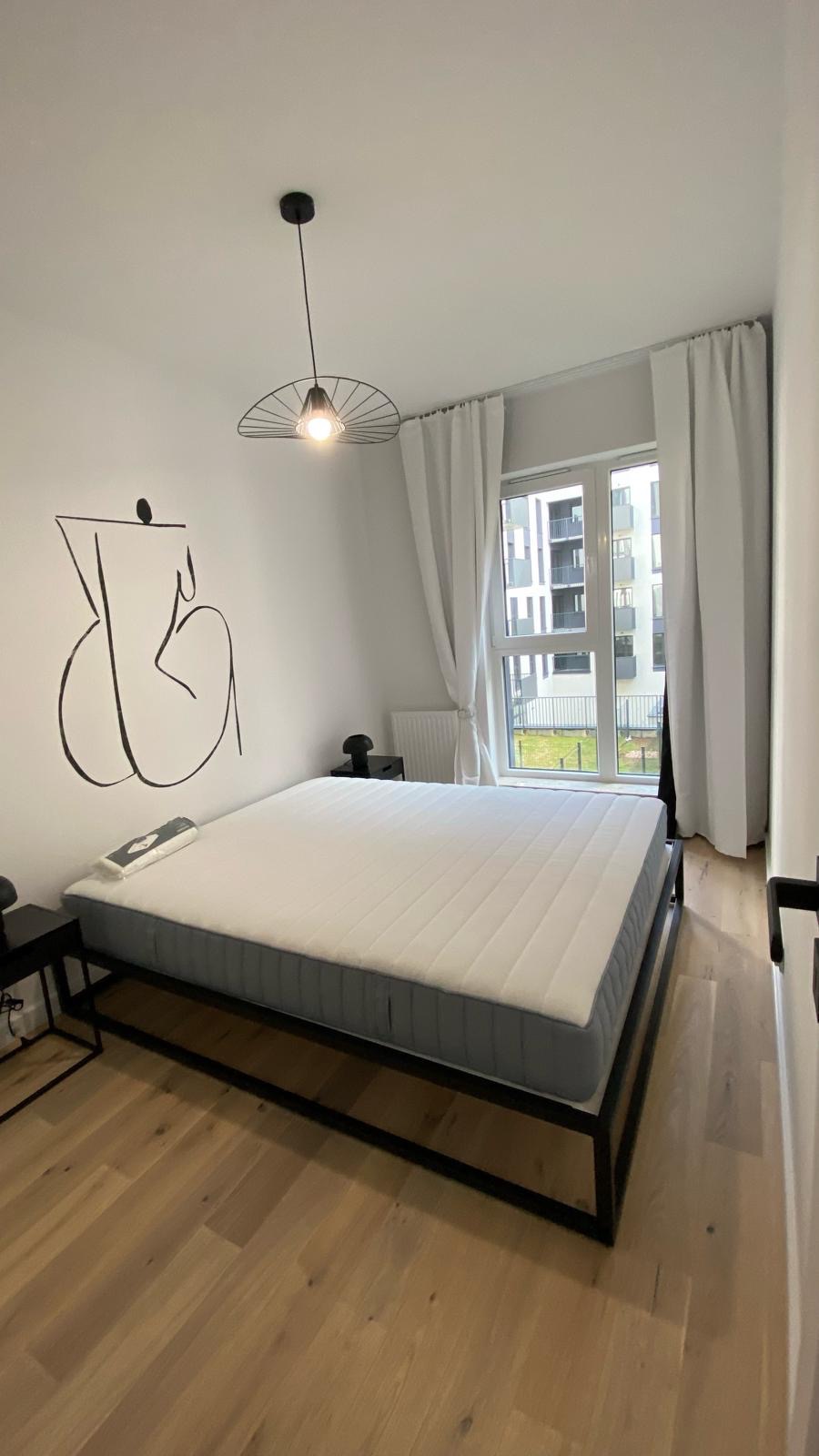 Minimalistyczna sypialnia z łóżkiem na czarnej ramie, drewnianą podłogą i abstrakcyjnym rysunkiem na ścianie, doświetlona przez wiszącą lampę i światło dzienne z okna.