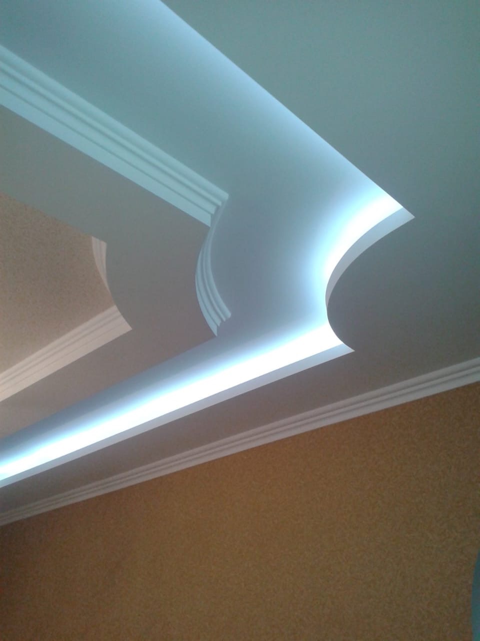 Podwieszany sufit z oświetleniem LED, dekoracyjne listwy przysufitowe i fragment ściany z tapetą o strukturze piasku.