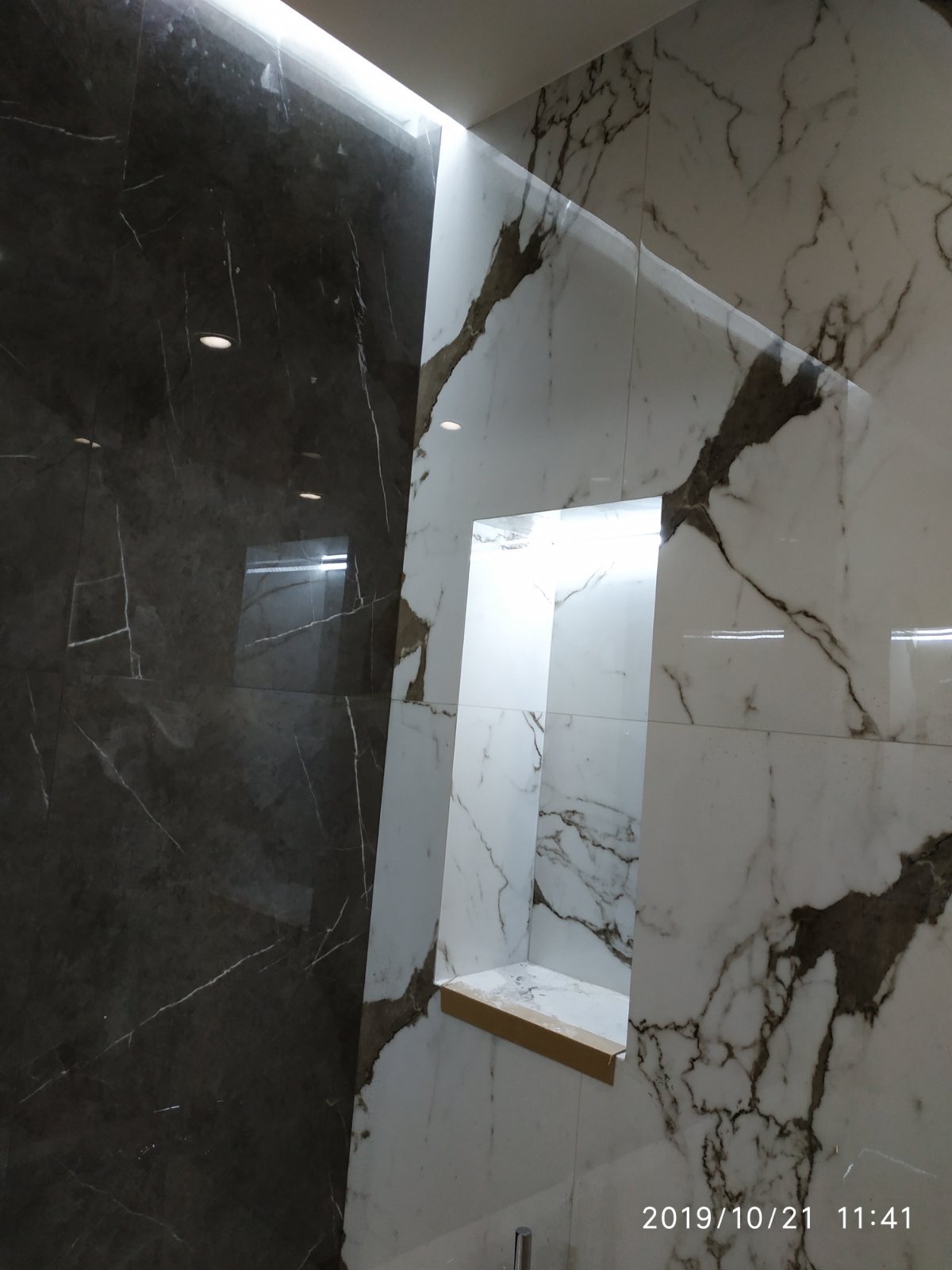 Wykończona wnęka w ścianie łazienki, obłożona białymi płytkami imitującymi marmur z ciemnymi żyłami, sąsiadująca z ciemną, błyszczącą ścianą również z marmurowym wzorem, oświetlenie LED.