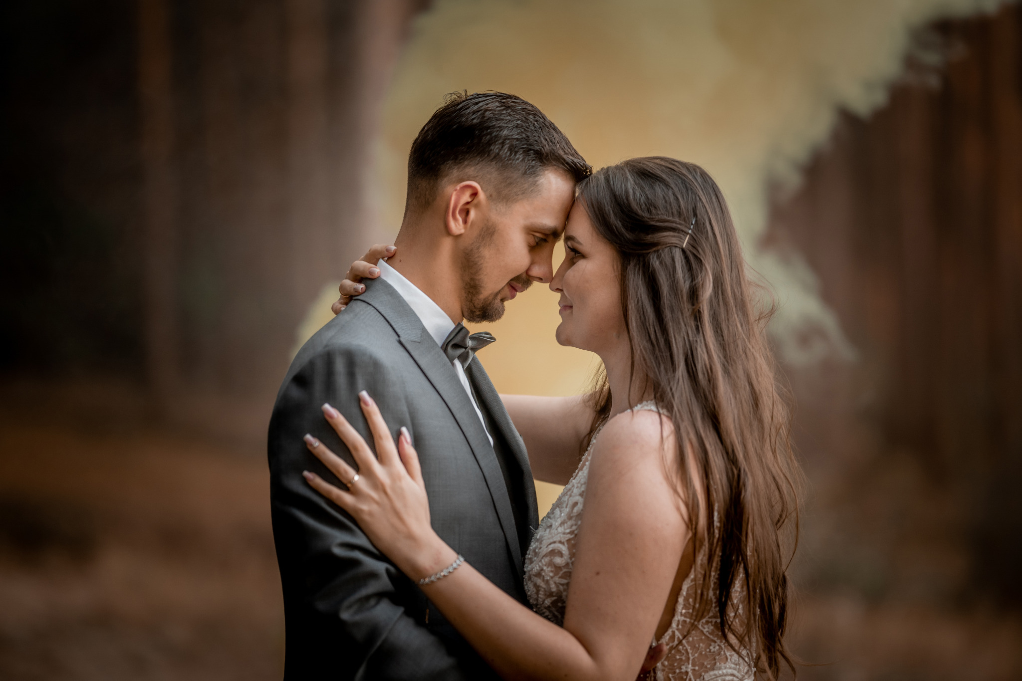 Nowożeńcy w zbliżeniu, dotykający się czołami na tle dymu, panna młoda w sukni z koronkowymi aplikacjami, pan młody w szarym garniturze z muchą.