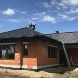 Dom w stanie surowym otwartym z ciemną dachówką, widoczne okna z roletami zewnętrznymi, mur z cegły ceramicznej, na dachu pracuje dekarz, błękitne niebo z chmurami.