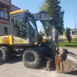 Żółto-czarna koparka Mecalac 12MTX45 na tle budynku mieszkalnego i drzew, z widocznym logo firmy KMRENT.PL na boku maszyny.