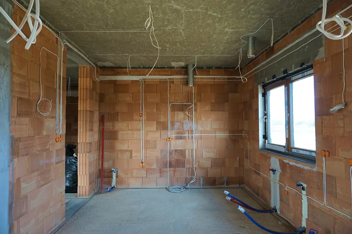 Wnętrze w stanie surowym z widoczną instalacją elektryczną i rurami grzewczymi, ściany z czerwonej cegły dziurawki, przewody poprowadzone po ścianach i suficie, puszki elektryczne zamontowane.