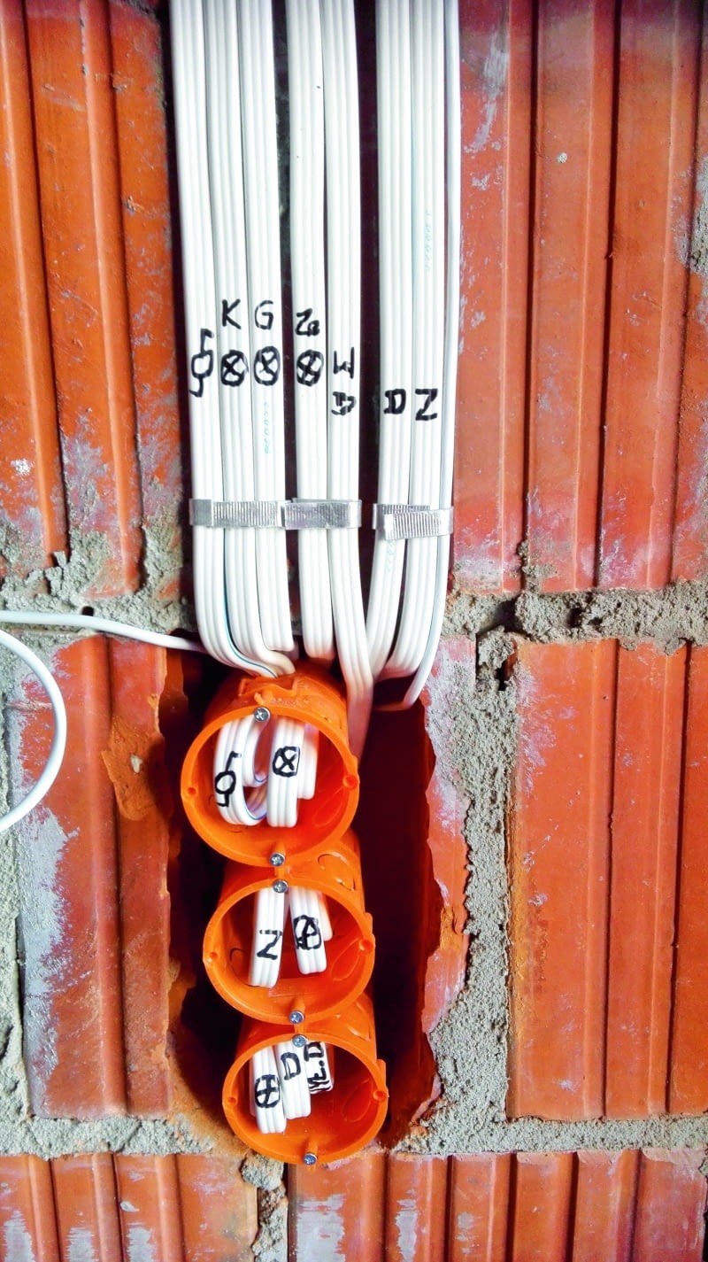 Białe przewody elektryczne z oznaczeniami, prowadzone w pomarańczowych puszkach instalacyjnych, zamontowane w ścianie z czerwonej cegły.