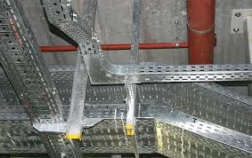 Metalowe korytka kablowe, mocowane do sufitu, widoczne przewody w kolorze czerwonym. System prowadzenia kabli w obiekcie przemysłowym.