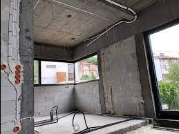 Surowy stan wnętrza z widocznymi przewodami elektrycznymi prowadzonymi po suficie i ścianach, puszki elektryczne zamontowane na ścianie z betonu komórkowego, okna bez parapetów.