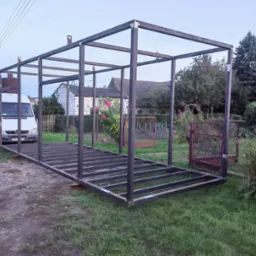 Surowa, spawana konstrukcja szkieletowa z metalowych profili na trawie, z białym busem w tle, widoczne spawy i niedoskonałości wykończenia.