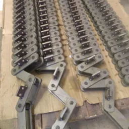 Rzędy metalowych łańcuchów transportowych z przyspawanymi elementami, ułożone na kartonowym podłożu, widoczne ślady spawania.