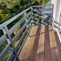 Szara balustrada nierdzewna na balkonie z deskami kompozytowymi, widok na zieleń drzew i błękitne niebo.