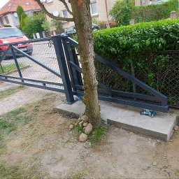 Prodan - Nowa, przesuwna brama wjazdowa z metalową konstrukcją, zamontowana na betonowej podstawie obok drzewa, z widocznym fragmentem ogrodzenia z siatki, leżącym psem i osobą w czerwonym samochodzie w tle.