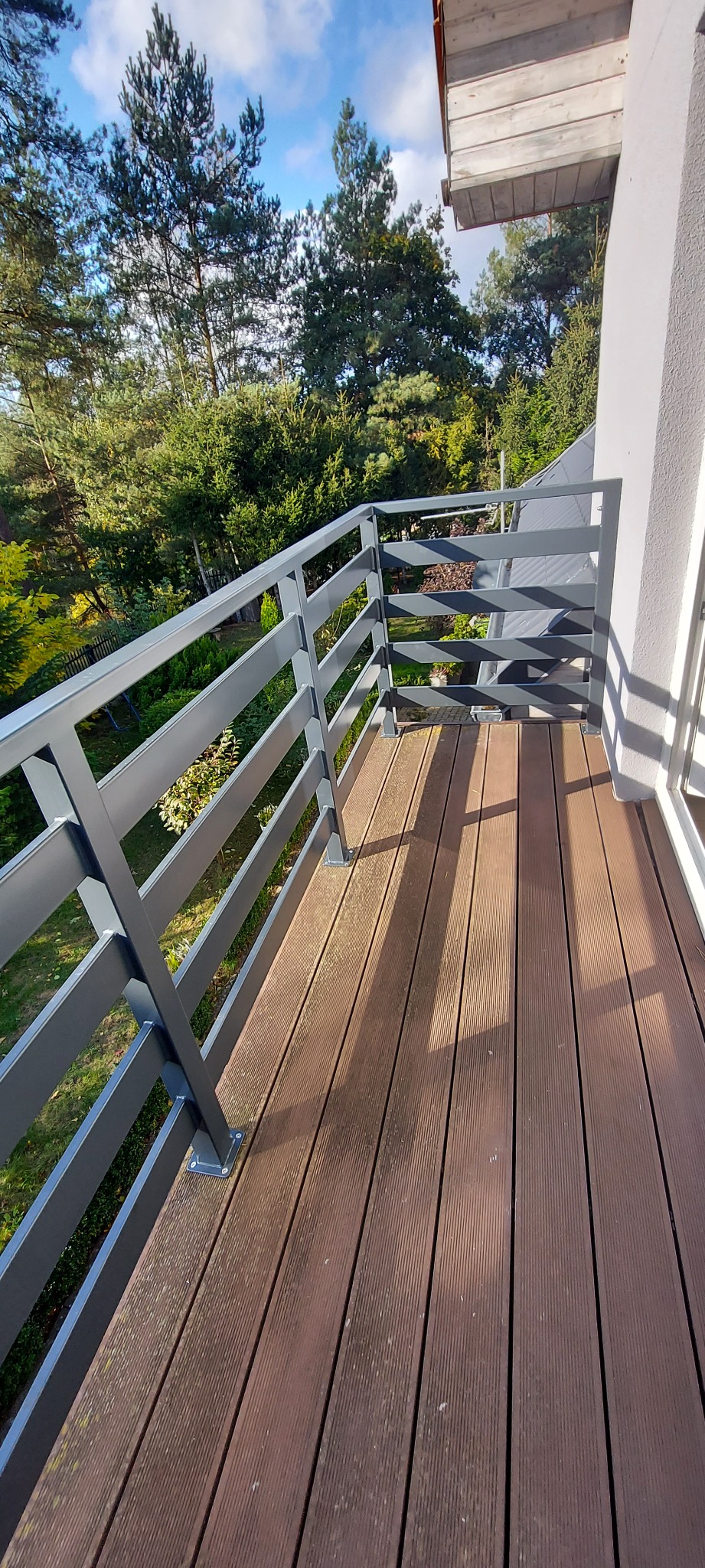 Szara balustrada nierdzewna na balkonie z deskami kompozytowymi, widok na zieleń drzew i błękitne niebo.