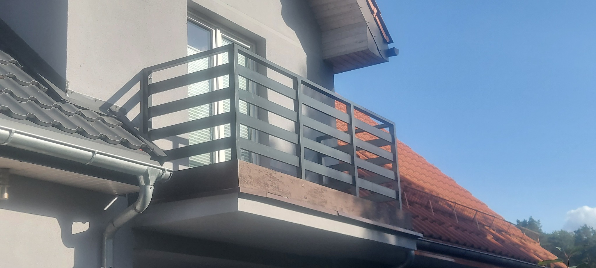 Szara, pozioma balustrada balkonowa z metalowych profili, widok od dołu na tle błękitnego nieba i dachu sąsiedniego budynku.