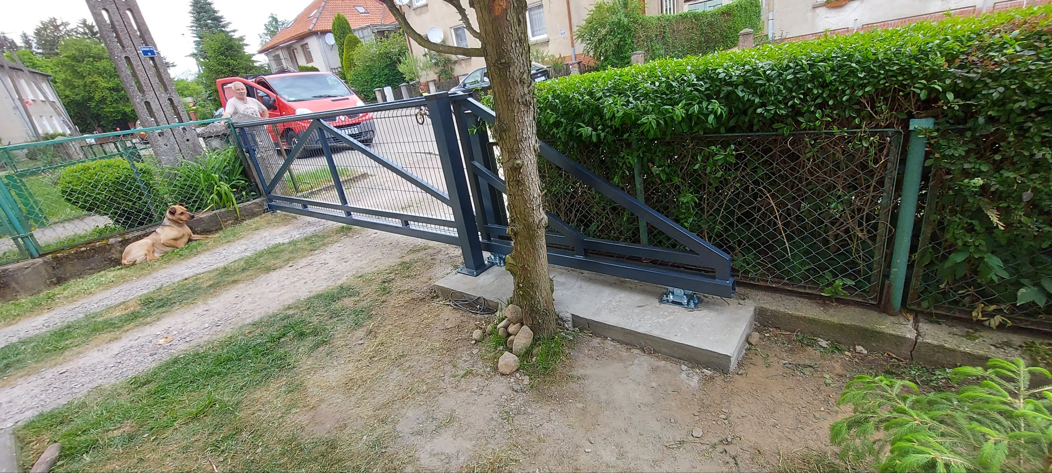 Nowa, przesuwna brama wjazdowa z metalową konstrukcją, zamontowana na betonowej podstawie obok drzewa, z widocznym fragmentem ogrodzenia z siatki, leżącym psem i osobą w czerwonym samochodzie w tle.