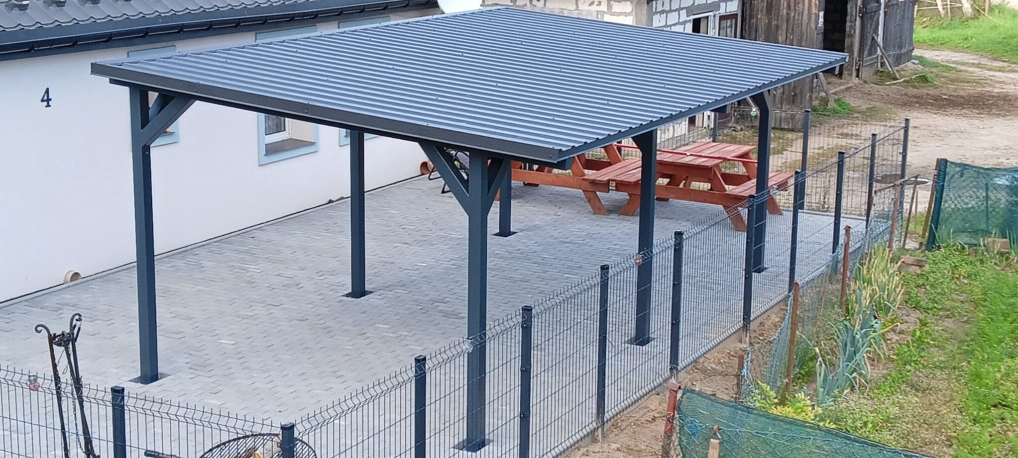 Zadaszona wiata z metalową konstrukcją i falistym dachem, obok drewniany stół piknikowy, całość otoczona ogrodzeniem panelowym.