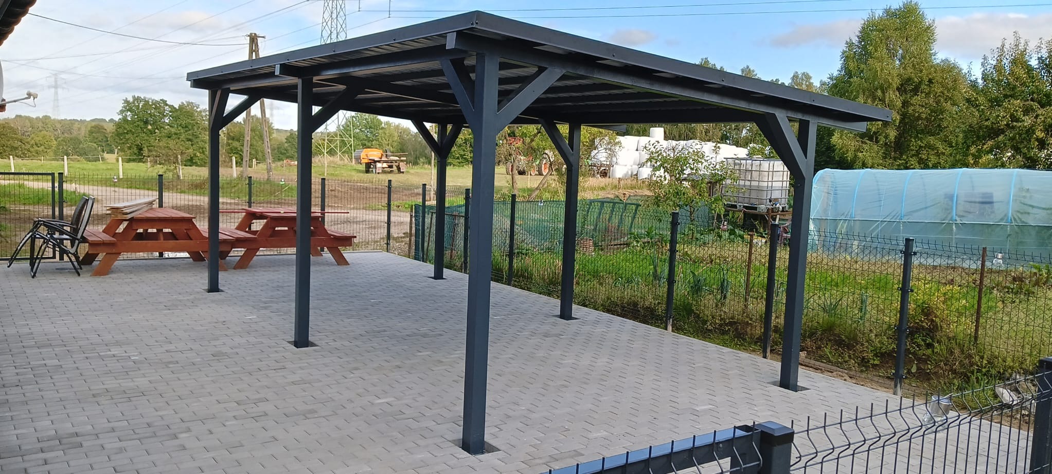 Zadaszona wiata ogrodowa z drewnianymi stołami piknikowymi, widok na ogród z tunelem foliowym i ogrodzeniem z siatki.