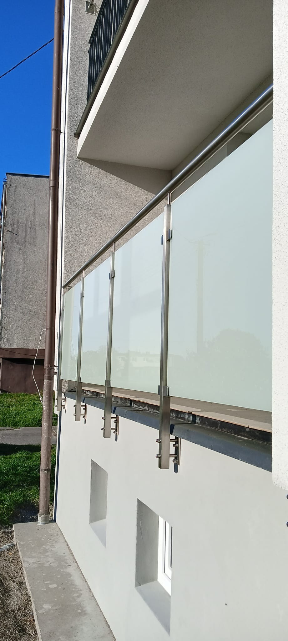 Nowoczesna balustrada balkonowa z matowego szkła i nierdzewnej stali, zamocowana do ściany budynku z widocznymi elementami mocującymi.