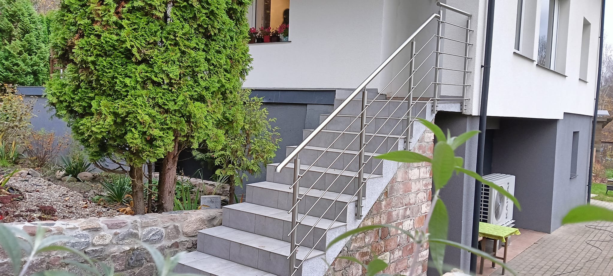 Zewnętrzne schody z szarych płytek prowadzące do budynku, zabezpieczone nowoczesną balustradą ze stali nierdzewnej z poziomymi poprzeczkami, otoczone zielenią i kamiennym murkiem.