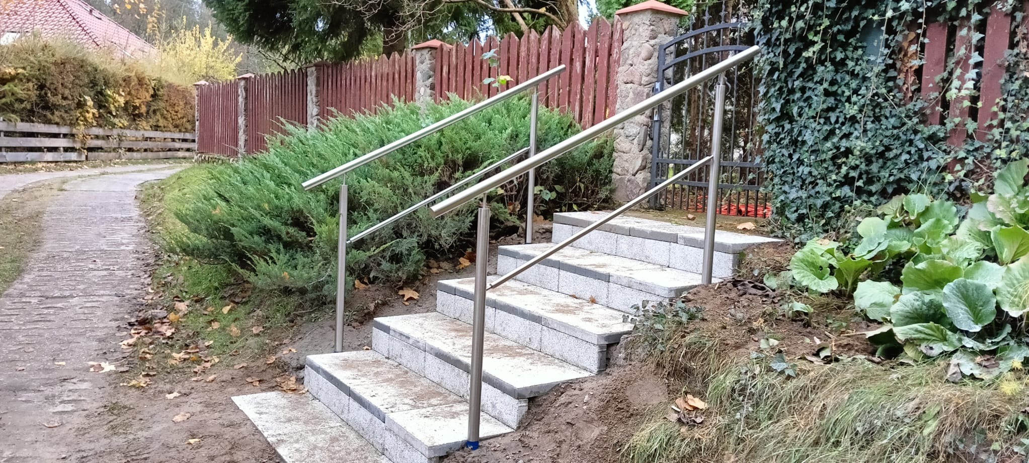 Zewnętrzne schody z jasnoszarej kostki brukowej z poręczą ze stali nierdzewnej, prowadzące do furtki w drewnianym ogrodzeniu, otoczone zielenią.