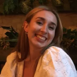 Portret uśmiechniętej młodej kobiety z blond włosami, w białej bluzce z ażurowymi detalami, na tle roślin i ceglanej ściany.